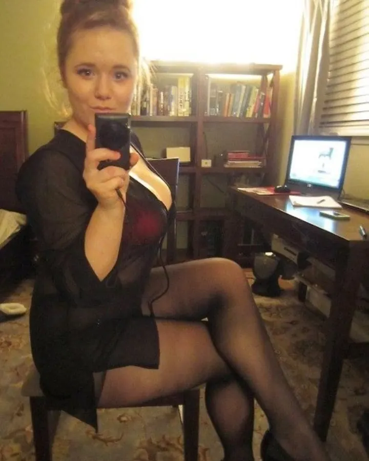 Jeune femme blonde en robe noire transparente, tenant un téléphone, assise sur une chaise dans une pièce avec un bureau et un ordinateur.