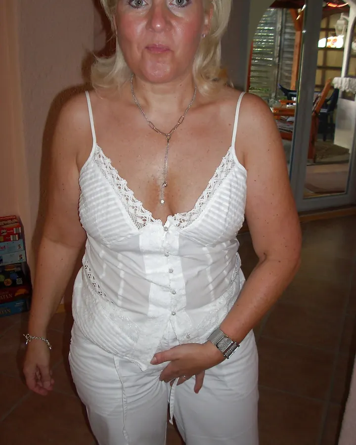 Femme blonde en haut blanc à lacets, posant debout dans une pièce, mains sur les hanches.