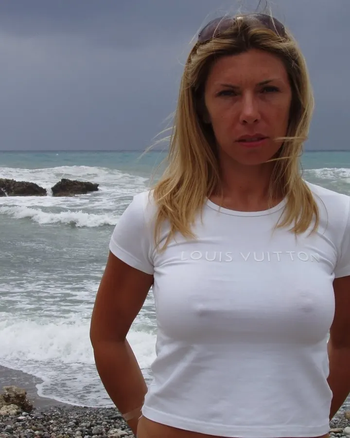 Femme blonde en t-shirt blanc Louis Vuitton, debout sur une plage rocheuse, regardant l'appareil photo