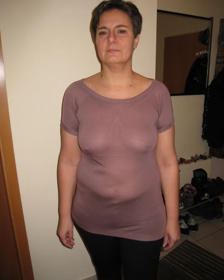 Femme debout, portant un t-shirt mauve à col rond, pantalon noir, regardant l'appareil, devant un mur beige.