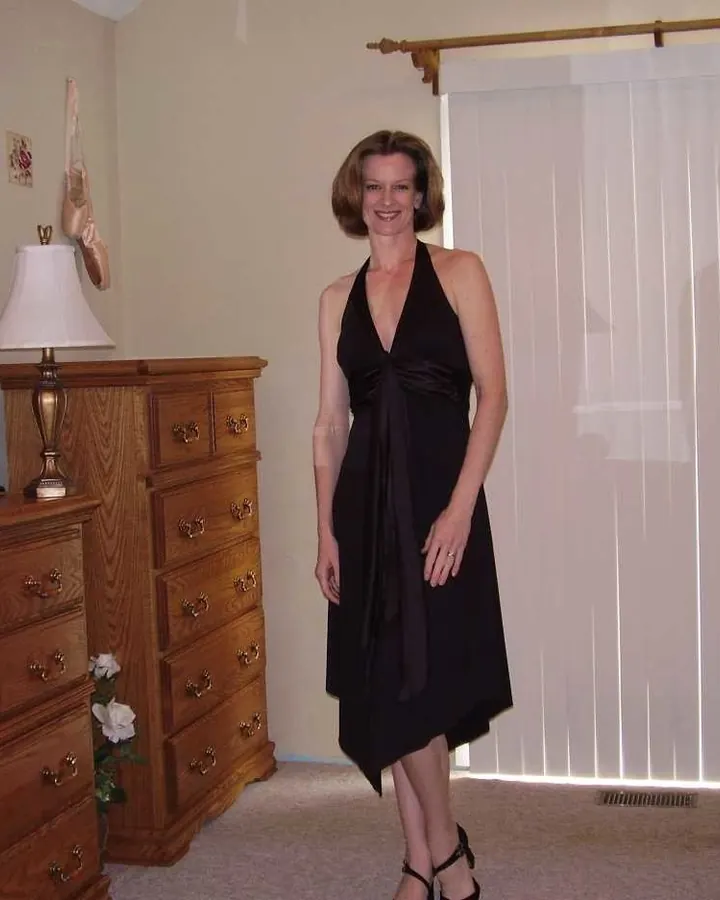 Femme aux cheveux châtains portant une robe noire, souriante dans une chambre avec un meuble en bois et des rideaux blancs.