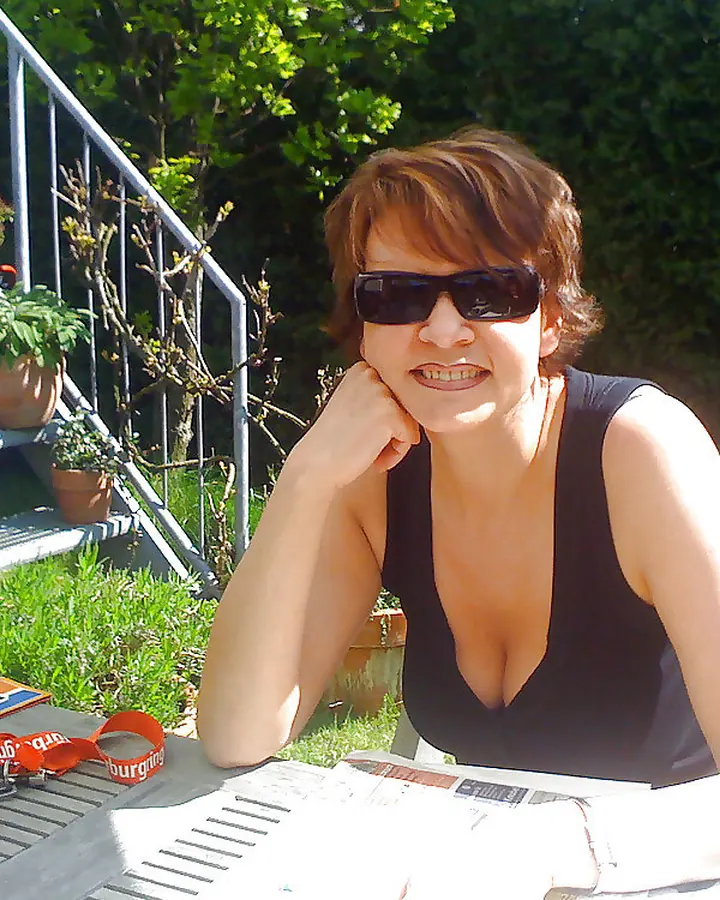 Femme aux cheveux roux, en haut noir, assise à une table en extérieur, portant des lunettes de soleil, regardant vers l'appareil.
