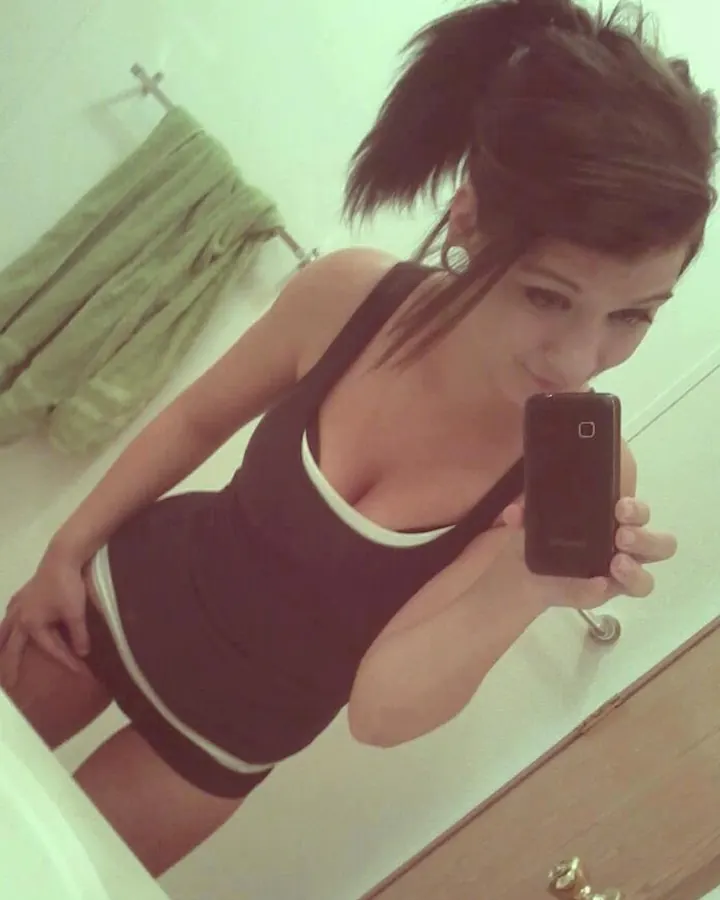 Femme aux cheveux bruns en tenue sombre, prenant un selfie dans un miroir de salle de bain, souriant légèrement.