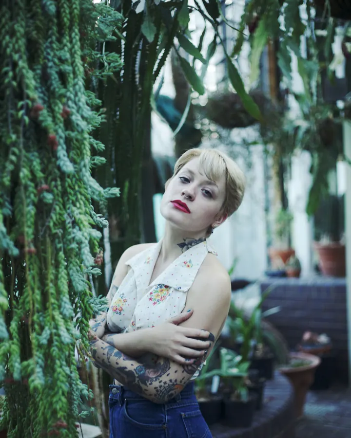 Femme aux cheveux blonds courts, vêtue d'un haut blanc à motifs floraux, jeans, tatouages visibles, bras croisés, regardant l'appareil, entourée de plantes.