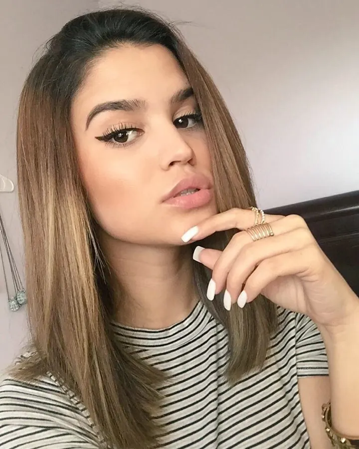 Jeune femme aux cheveux châtains, portant un haut rayé noir et blanc, regardant l'appareil avec un doigt sur les lèvres, maquillage soigné.