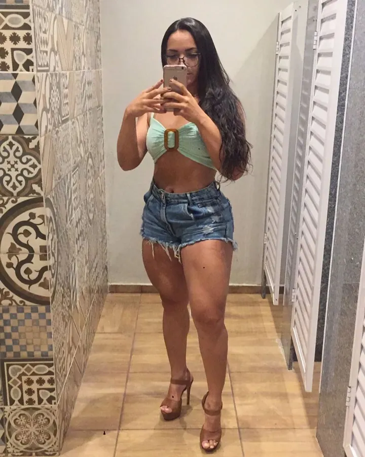 Femme aux cheveux longs bruns foncés, portant un haut vert menthe avec boucle dorée et un short en jean, prenant un selfie devant un miroir, pieds chaussés de sandales à talons.