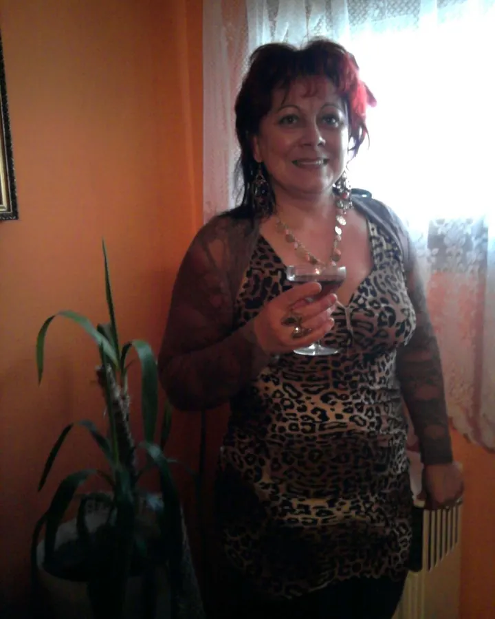 Femme aux cheveux roux, portant un haut à motif léopard et une veste transparente, tenant un verre de vin rouge, souriant devant une fenêtre.