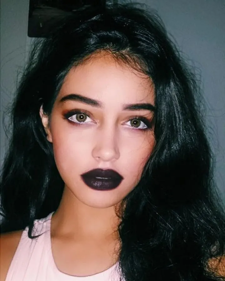 Jeune femme aux cheveux bruns foncés, maquillage prononcé avec rouge à lèvres sombre, regardant directement l'appareil photo.