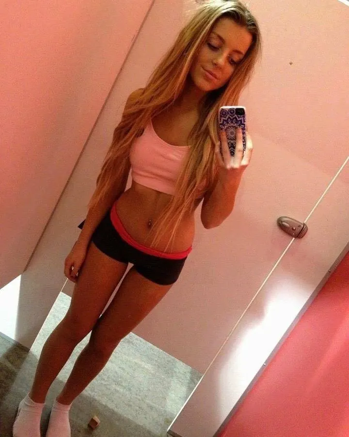 Jeune femme blonde prenant un selfie devant un miroir, portant un haut rose et un short noir avec bordure rose, tenant un téléphone à motifs.