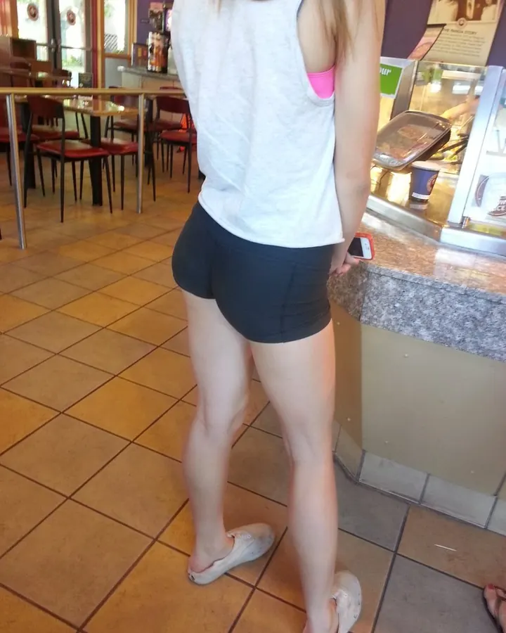 Vue arrière d'une jeune femme aux cheveux blonds, portant un haut blanc et un short noir, debout dans un café.