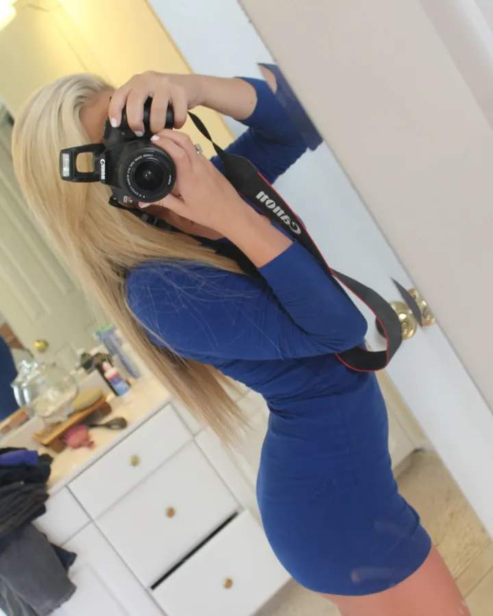 Jeune femme blonde, en robe bleue ajustée, prenant un selfie avec un appareil photo Canon dans une salle de bain.