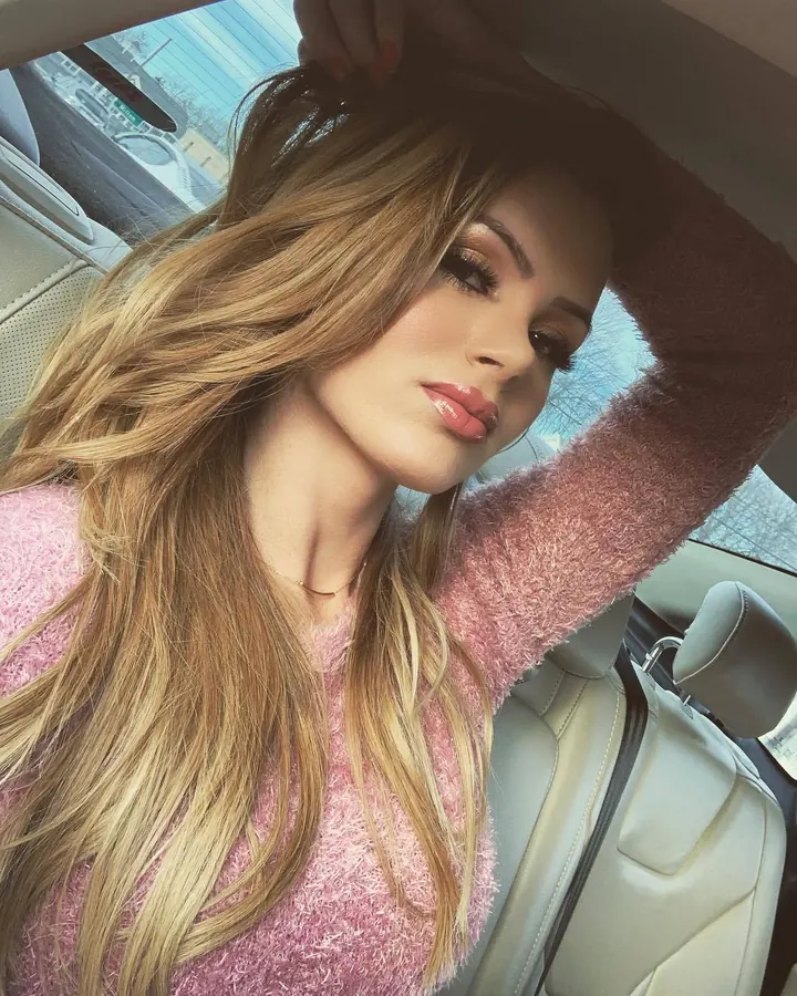 Femme blonde en pull rose, assise dans une voiture, touchant ses cheveux.