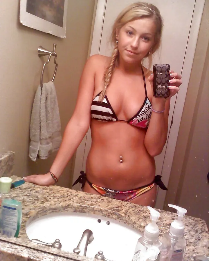 Jeune femme blonde aux cheveux tressés, prenant un selfie dans un miroir de salle de bain, portant un bikini multicolore, avec un piercing au nombril.