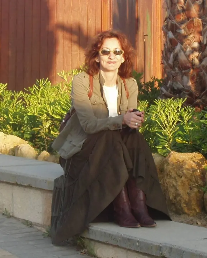Femme aux cheveux roux, portant des lunettes de soleil, assise sur un mur de pierre devant un bâtiment en bois, avec un sac à dos et des bottes marron.