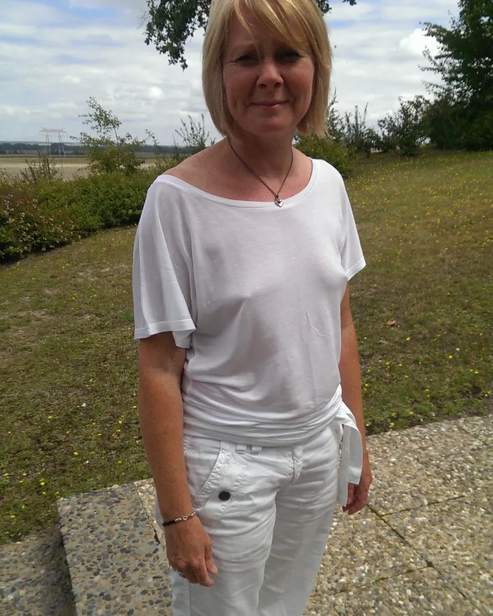 Femme blonde aux cheveux courts, vêtue d'un haut blanc et de pantalons blancs, debout dans un champ verdoyant sous un ciel nuageux.
