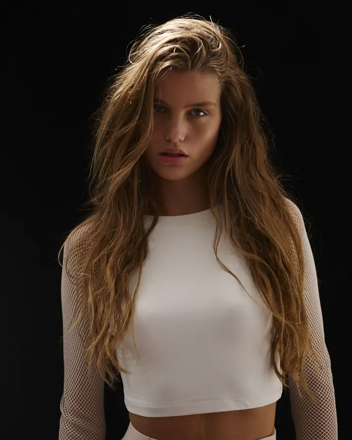 Femme aux cheveux châtains en haut blanc et maille fine posant devant un fond noir, regardant l'appareil