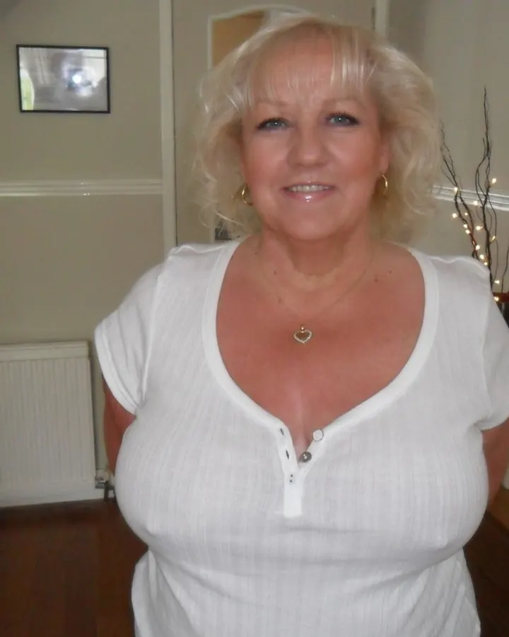 Femme blonde aux cheveux courts, en chemisier blanc, souriant dans une pièce intérieure avec un radiateur et des décorations.