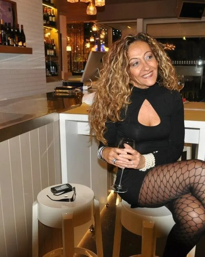 Femme aux cheveux blonds mi-longs, assise à un bar, portant une robe noire à découpe au décolleté, des collants noirs à mailles et tenant un verre de vin.