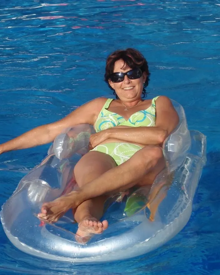 Femme aux cheveux bruns foncés, souriante, portant un maillot de bain vert et jaune, flottant sur un anneau gonflable dans une piscine bleue.