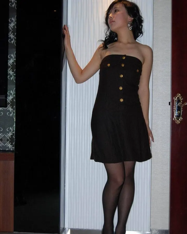 Jeune femme asiatique aux cheveux noirs, en robe noire à boutons dorés, posant de profil dans une pièce intérieure.
