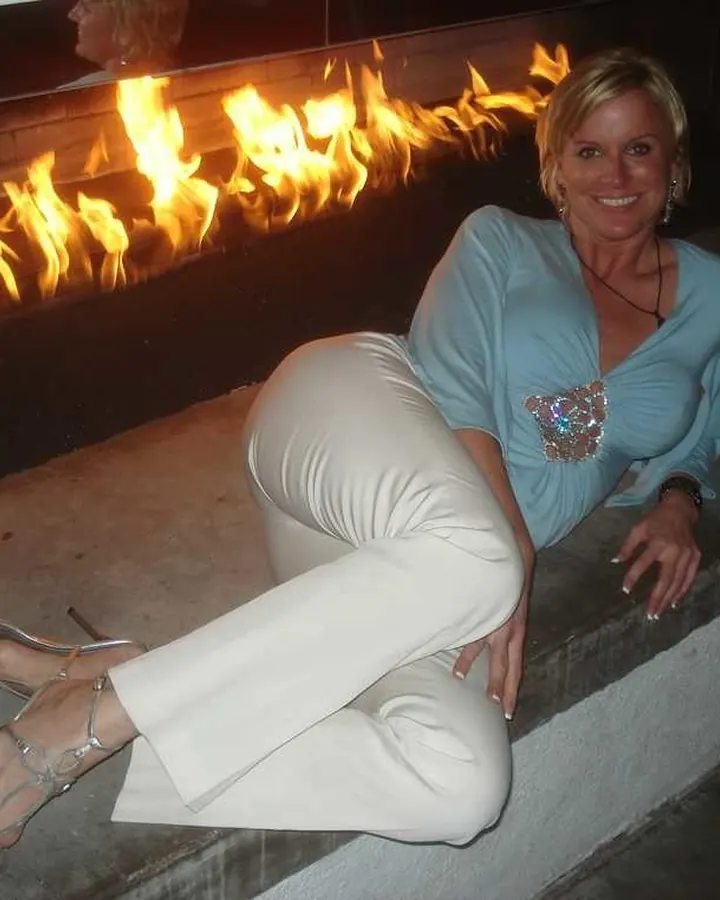 Femme mûre aux cheveux blonds, en chemisier bleu et pantalon blanc, assise devant une cheminée allumée.