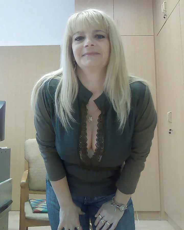 Femme blonde en chemise sombre, debout dans une pièce, mains sur les hanches