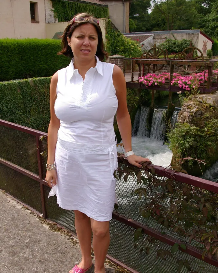 Femme aux cheveux châtains, en robe blanche, posant sur un pont en bois, regardant vers le bas, lunettes de soleil roses.