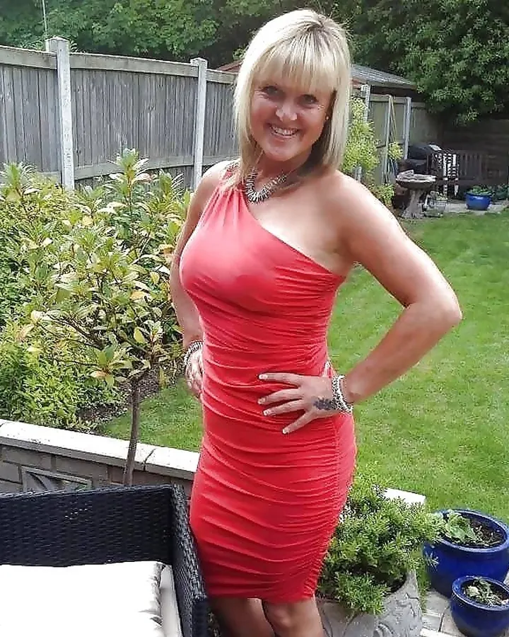 Femme en robe rouge ajustée, debout dans un jardin, souriant à la caméra avec une main sur la hanche.