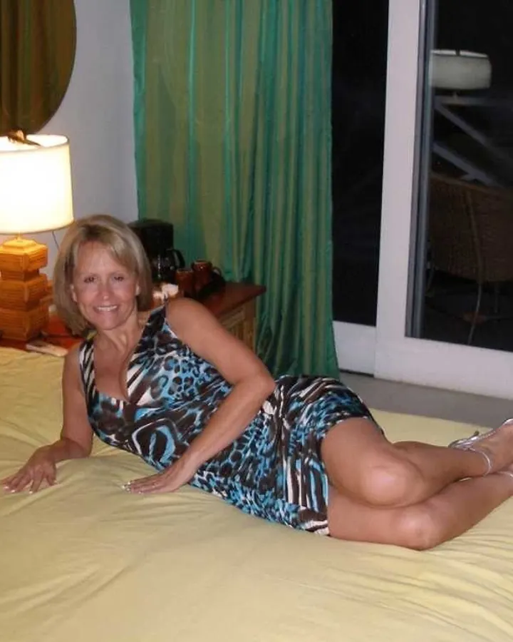 Femme blonde en robe bleue, allongée sur un lit jaune, regardant l’objectif.