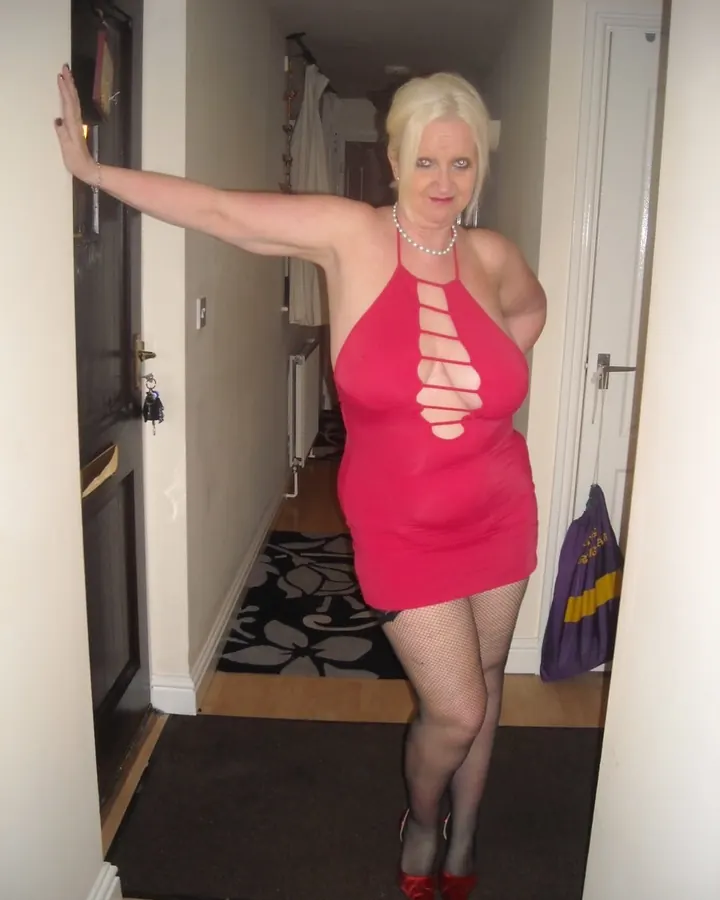 Femme blonde en haut noir transparent, robe noire, talons noirs, debout dans une chambre, mains croisées devant elle.