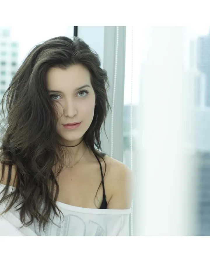Jeune femme aux cheveux bruns noirs, en débardeur blanc et soutien-gorge noir, regardant l'appareil, devant une fenêtre avec vue sur des gratte-ciel.