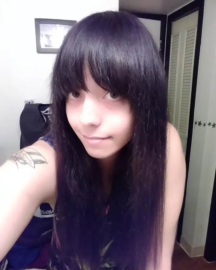 Femme en photo selfie, cheveux noirs longs, regardant l'appareil, tatouage visible sur l'épaule.