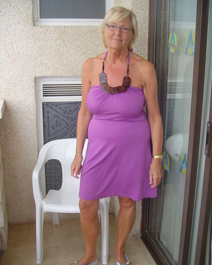 Femme blonde âgée portant une robe noire, des collants noirs et des escarpins, assise sur une chaise en bois dans une cuisine.