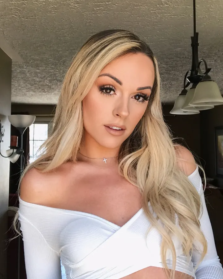 Femme blonde aux cheveux longs, maquillage naturel, vêtue d'un haut blanc décolleté, posant dans une pièce