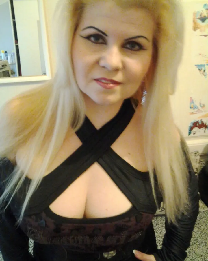 Femme blonde aux cheveux platine, maquillage marqué, vêtue d'un haut noir croisé, regardant l'appareil