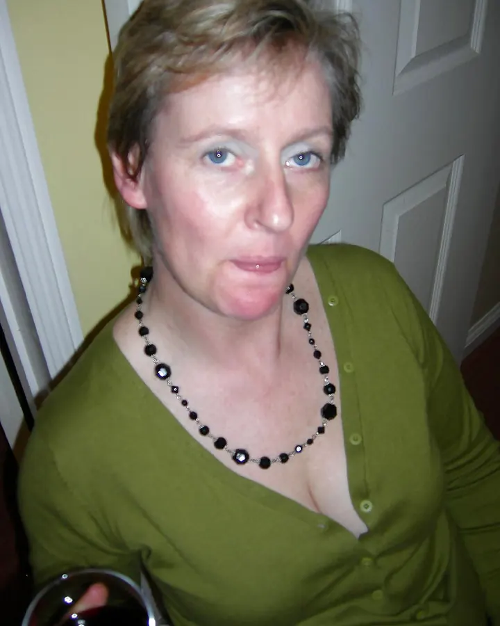 Femme européenne de 60 ans en intérieur, chemisier vert olive boutonné, col ouvert, cheveux courts blonds, regard direct, collier noir à perles.