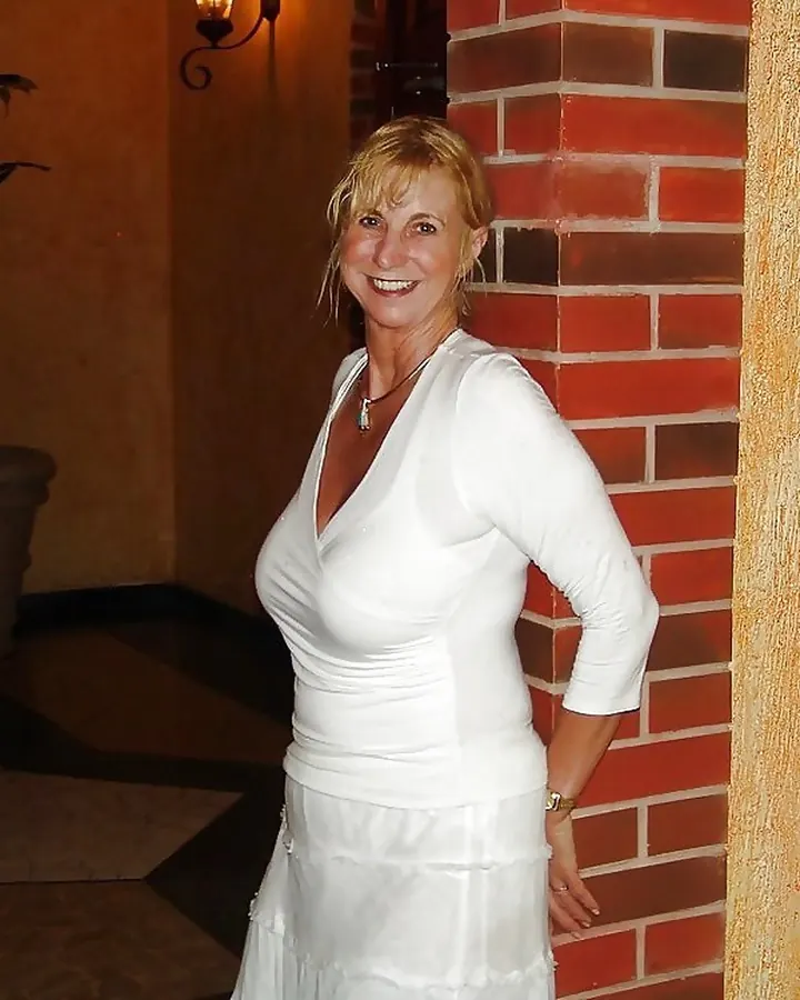 Femme blonde, en robe blanche, souriant, adossée à un mur de briques dans un hall intérieur