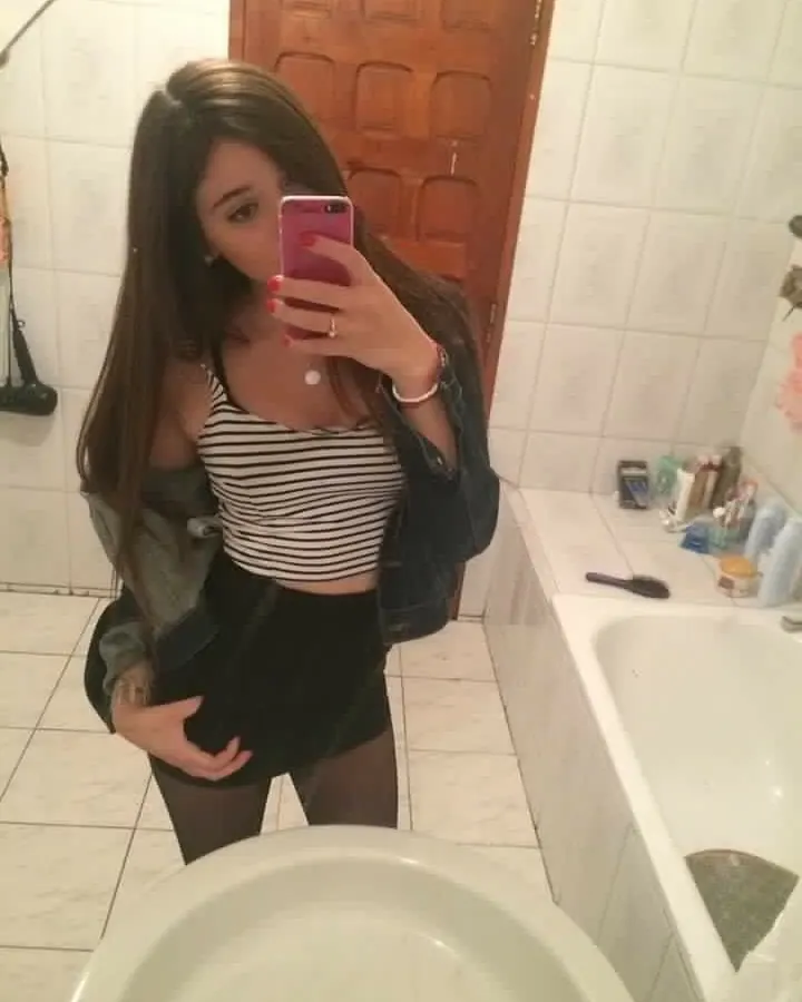 Femme aux cheveux bruns longs, en haut rayé noir-blanc et jupe noire, prenant un selfie devant un miroir dans une salle de bain