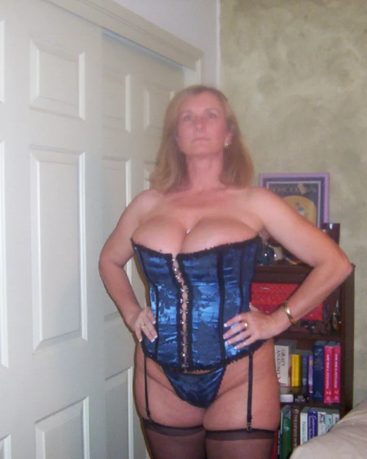 Femme en corset bleu métallisé, pose les mains sur les hanches dans une chambre.