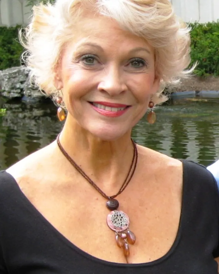 Femme aux cheveux blancs, en haut noir, souriante, devant un étang
