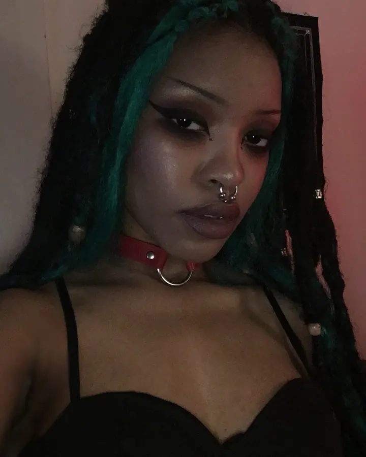 Femme aux cheveux noirs, coiffure tressée avec touches vertes, portant un collier rouge, regardant l'appareil photo