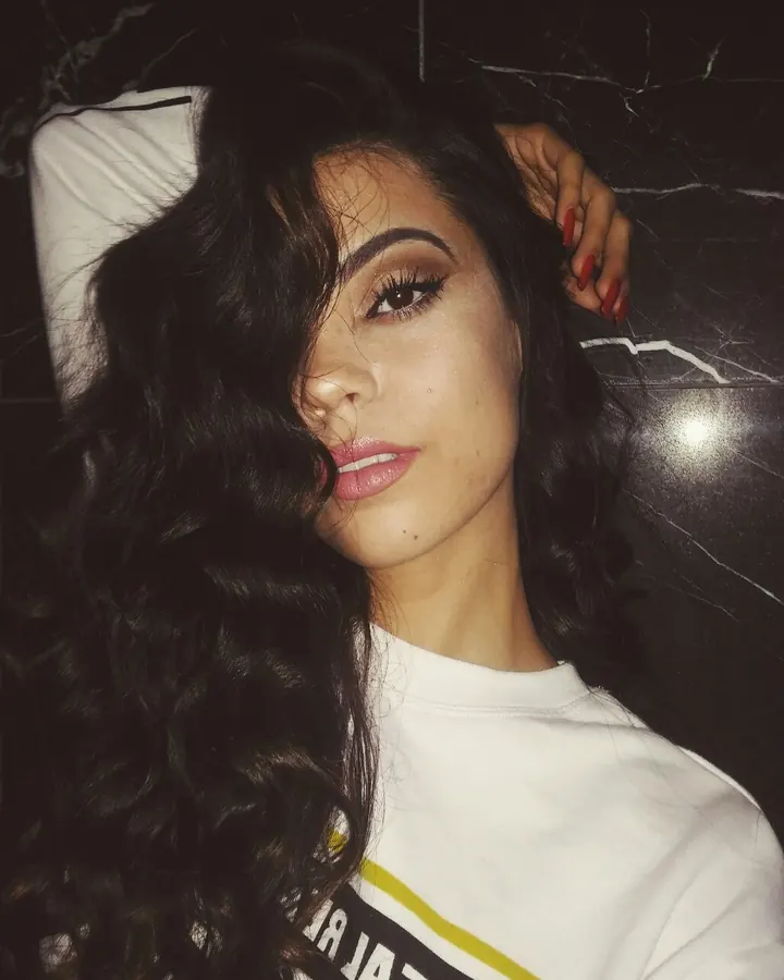 Femme aux cheveux noirs bouclés, maquillage prononcé, regardant l'objectif, portant un t-shirt blanc sur fond marbré noir