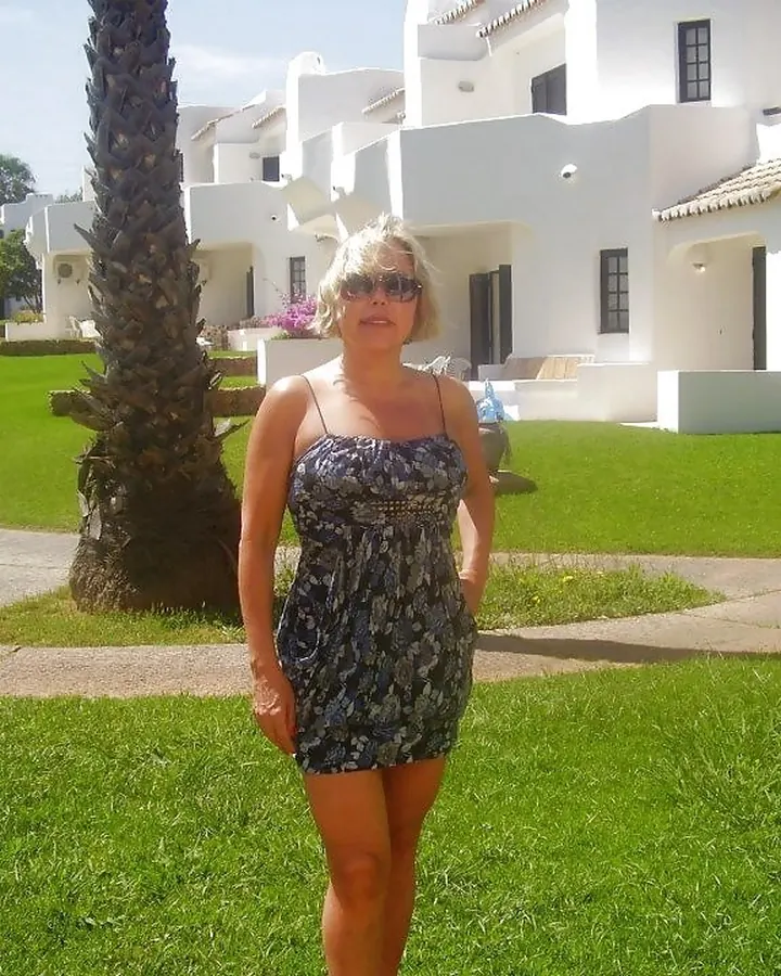 Femme blonde en robe imprimée, debout sur une pelouse verte devant des bâtiments blancs, lunettes de soleil.