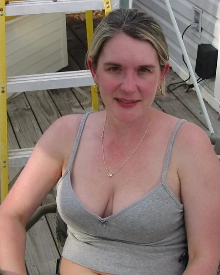 Femme aux cheveux blonds portant un haut gris, assise sur une chaise en bois sur une terrasse, regardant l'appareil.