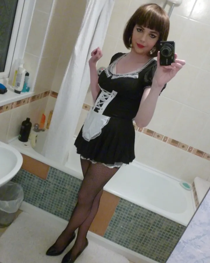 Femme aux cheveux châtains portant un haut noir et un maquillage foncé, prenant un selfie dans une salle de bain, tenant un appareil photo.