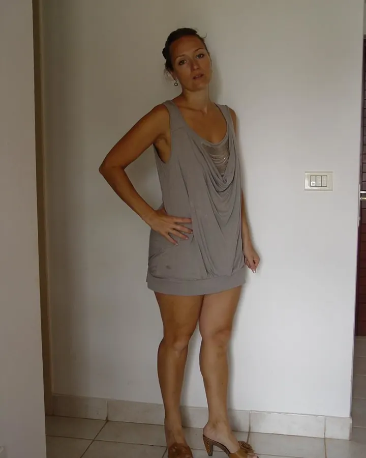 Femme aux cheveux bruns-noirs portant une robe grise, debout dans un couloir, main sur la hanche.