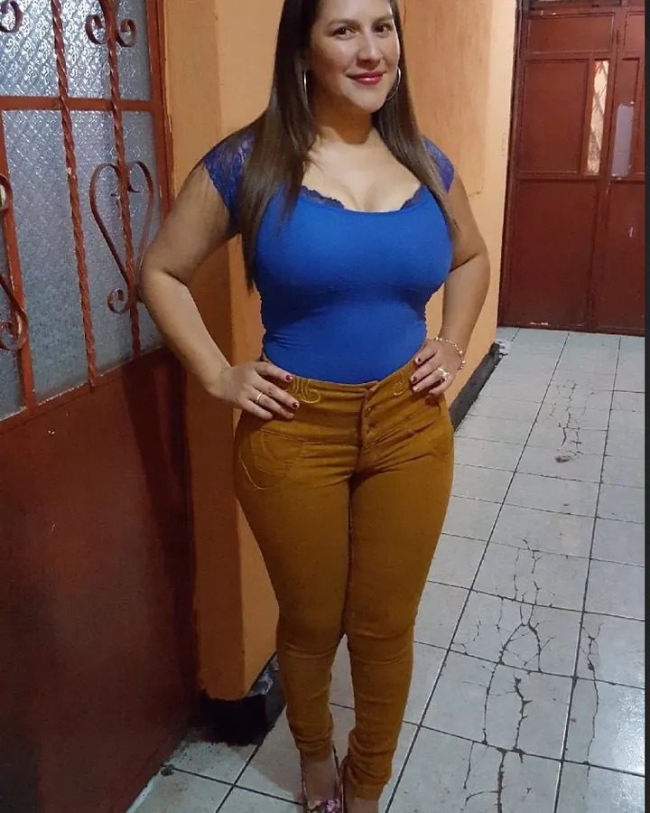 Femme en haut bleu et pantalon marron, souriante, les mains sur les hanches, dans un couloir aux carreaux fissurés.