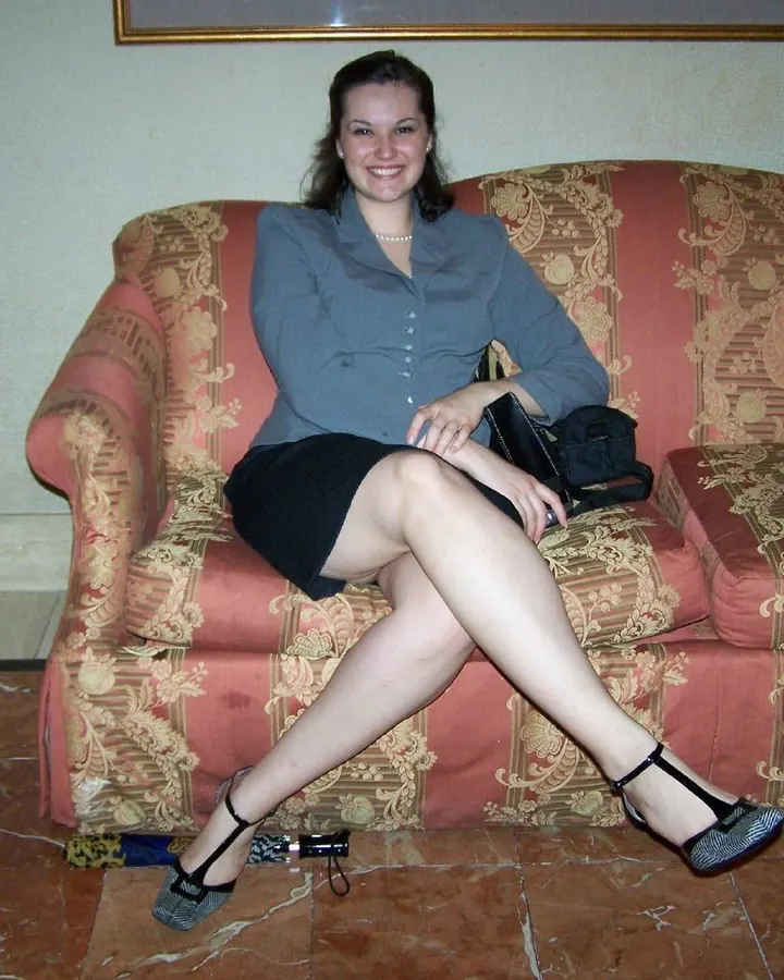 Femme blonde aux cheveux courts, en robe noire ajustée, assise sur un canapé rouge à motifs dorés, chaussures à talons noirs, main sur la cuisse, regardant l'appareil.
