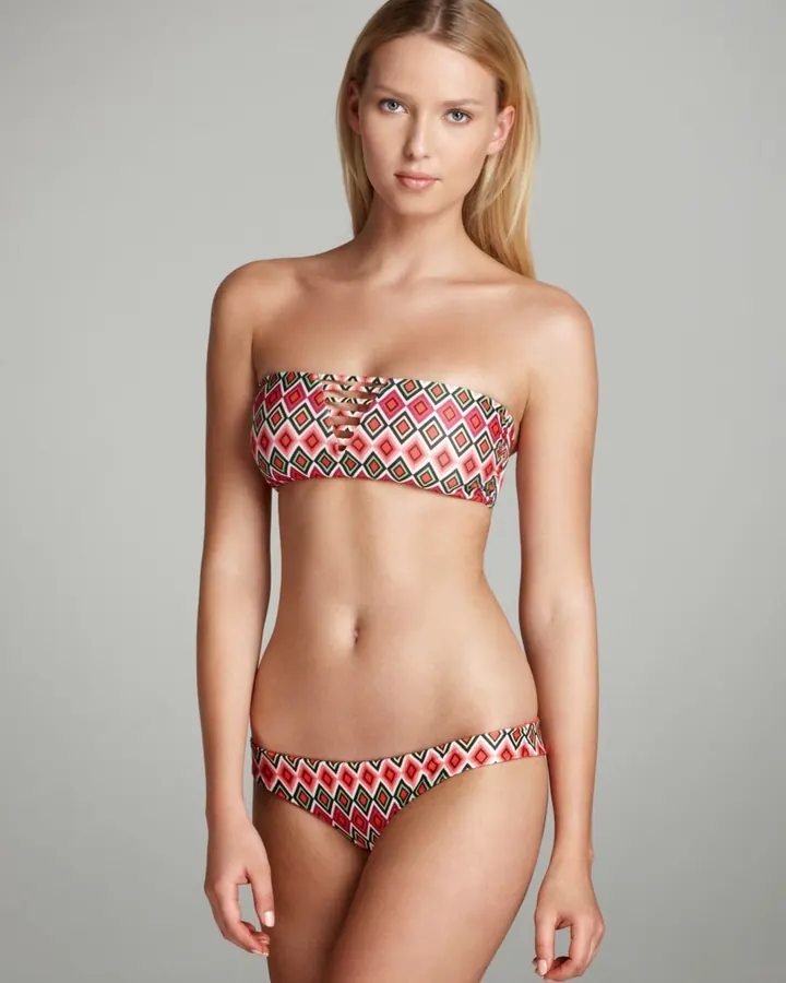 Femme blonde portant un bikini coloré à motifs géométriques, debout devant un fond gris neutre.