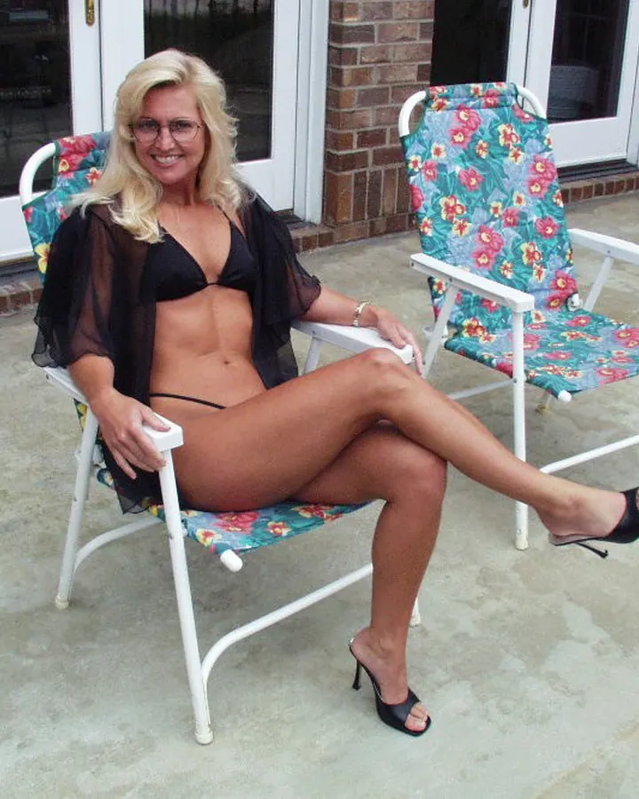 Femme aux cheveux blancs portant un maillot de bain noir, assise sur une chaise de jardin, souriant à la caméra.